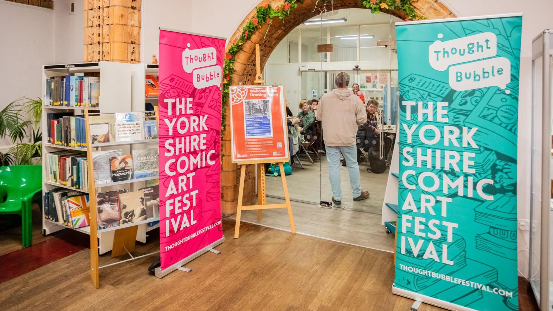 A library entrance is decorated by hanging flowers, with a red sign and a blue sign advertising Thought Bubble festival. The branding reads: "The Yorkshire comic art festival." A talk is going on in the background, with people looking at a speaker. 