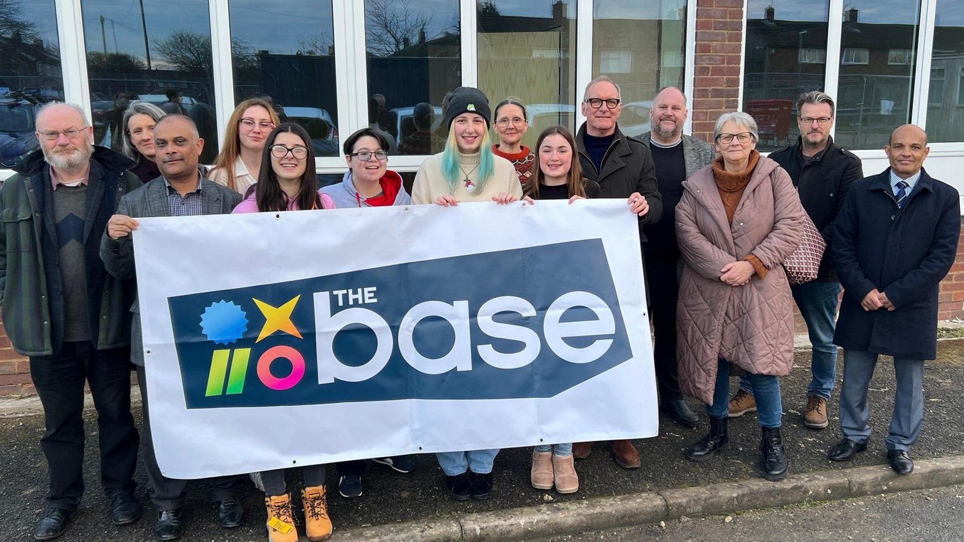 A group of people stood outside the building, which has eight windows and white panes. They are holding a banner that reads 'the base'.