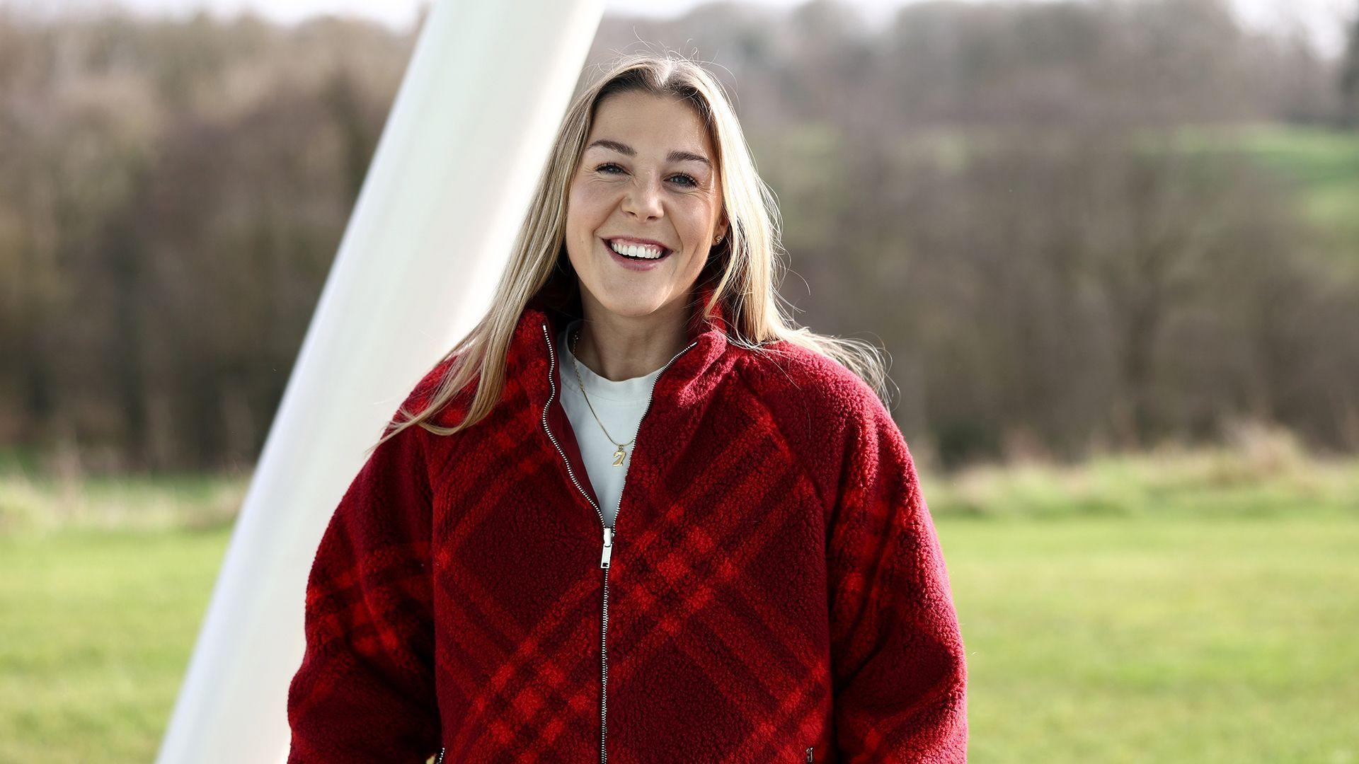 Mary Earps smiles while looking at the camera while standing outside against a backdrop of green grass and bare trees while wearing a red zip up top over a white shirt, at St. George's Park in February 2024.