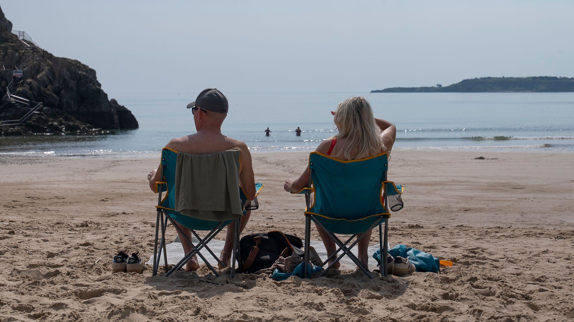 Two people on chair sitting on a beach. They are looking out at the sea. 