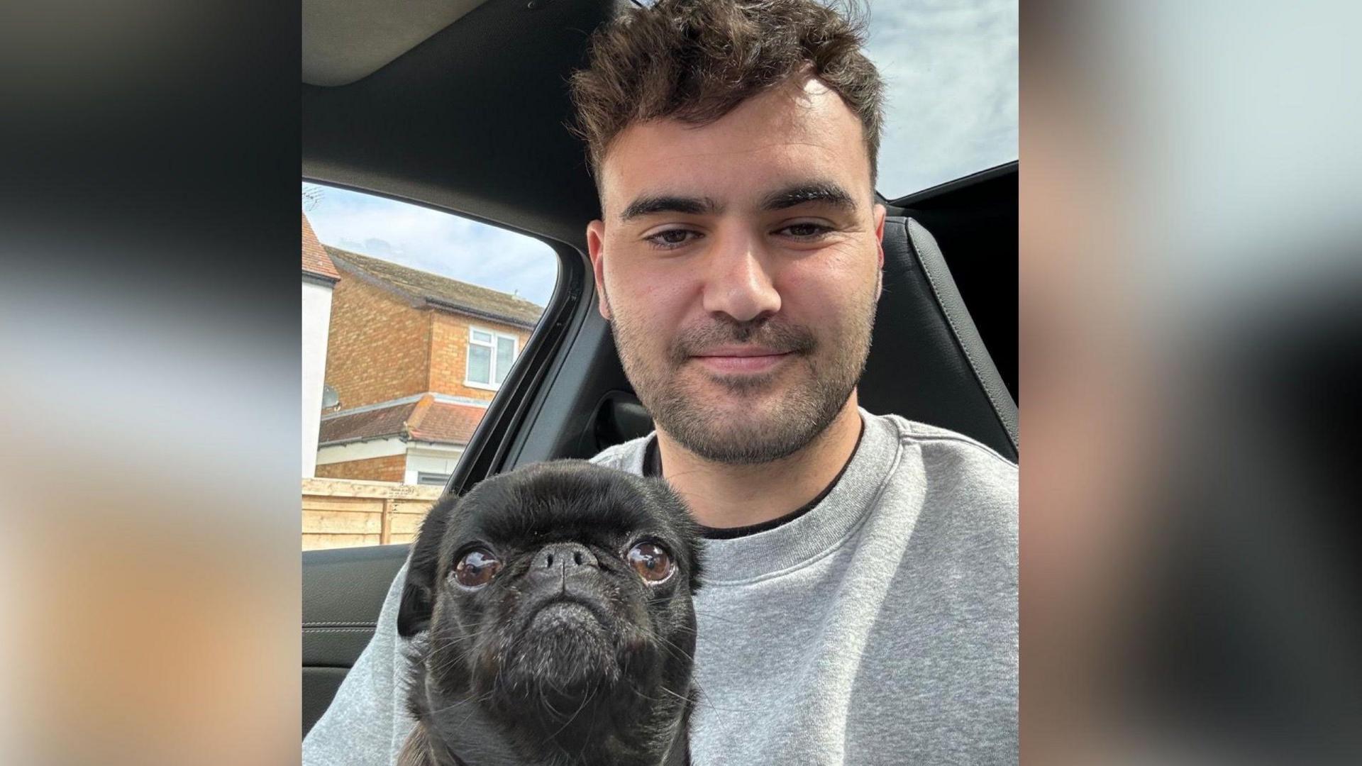 A young man, wearing a grey sweatshirt, sits in a car with a  small black pug dog on his lap. Both are looking towards the camera.
