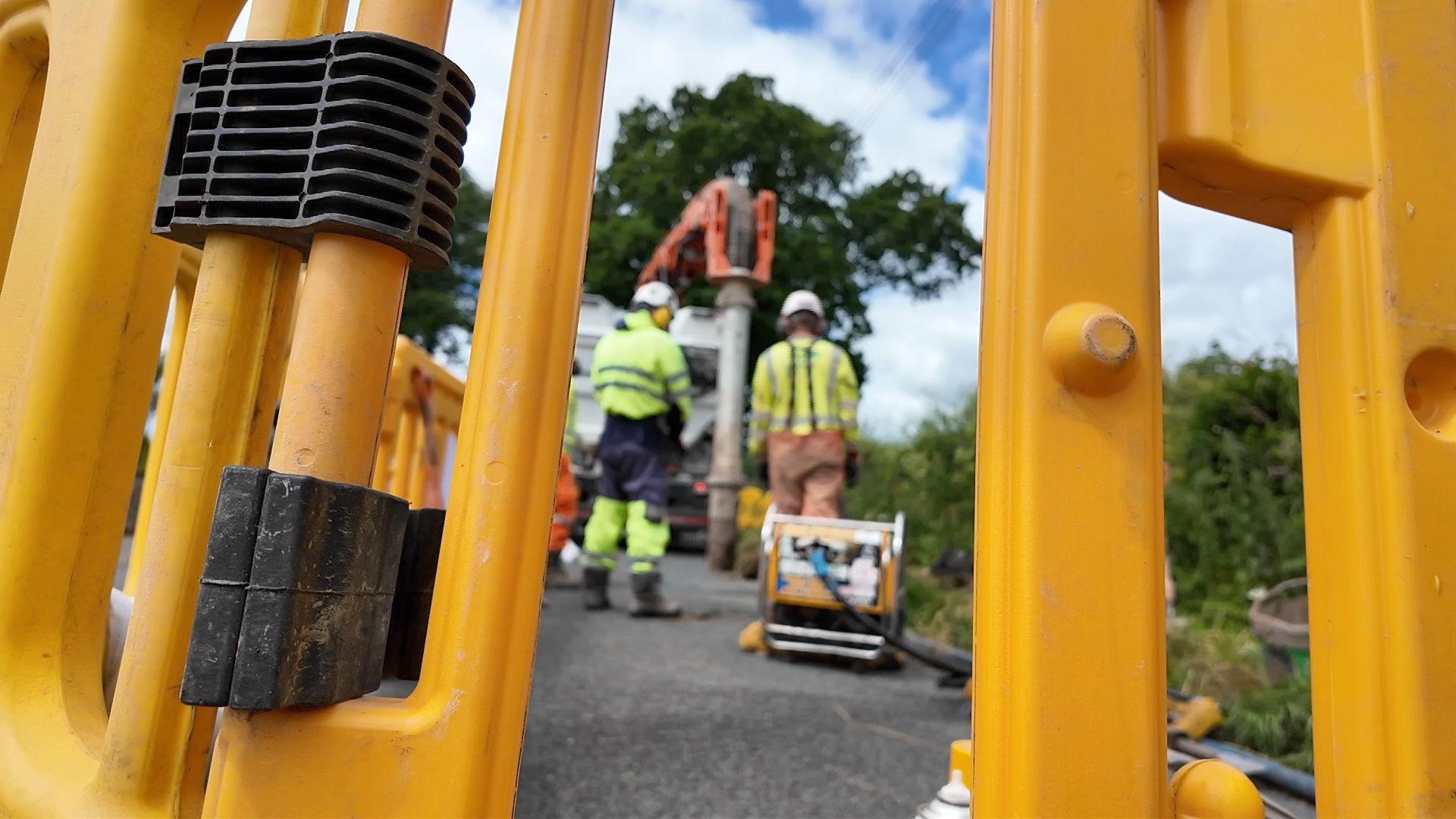 Staff are working to fix a leak at the side of a road in the countryside. The team are wearing high viz clothing and operating heavy machinery. The are working behind yellow plastic fencing.