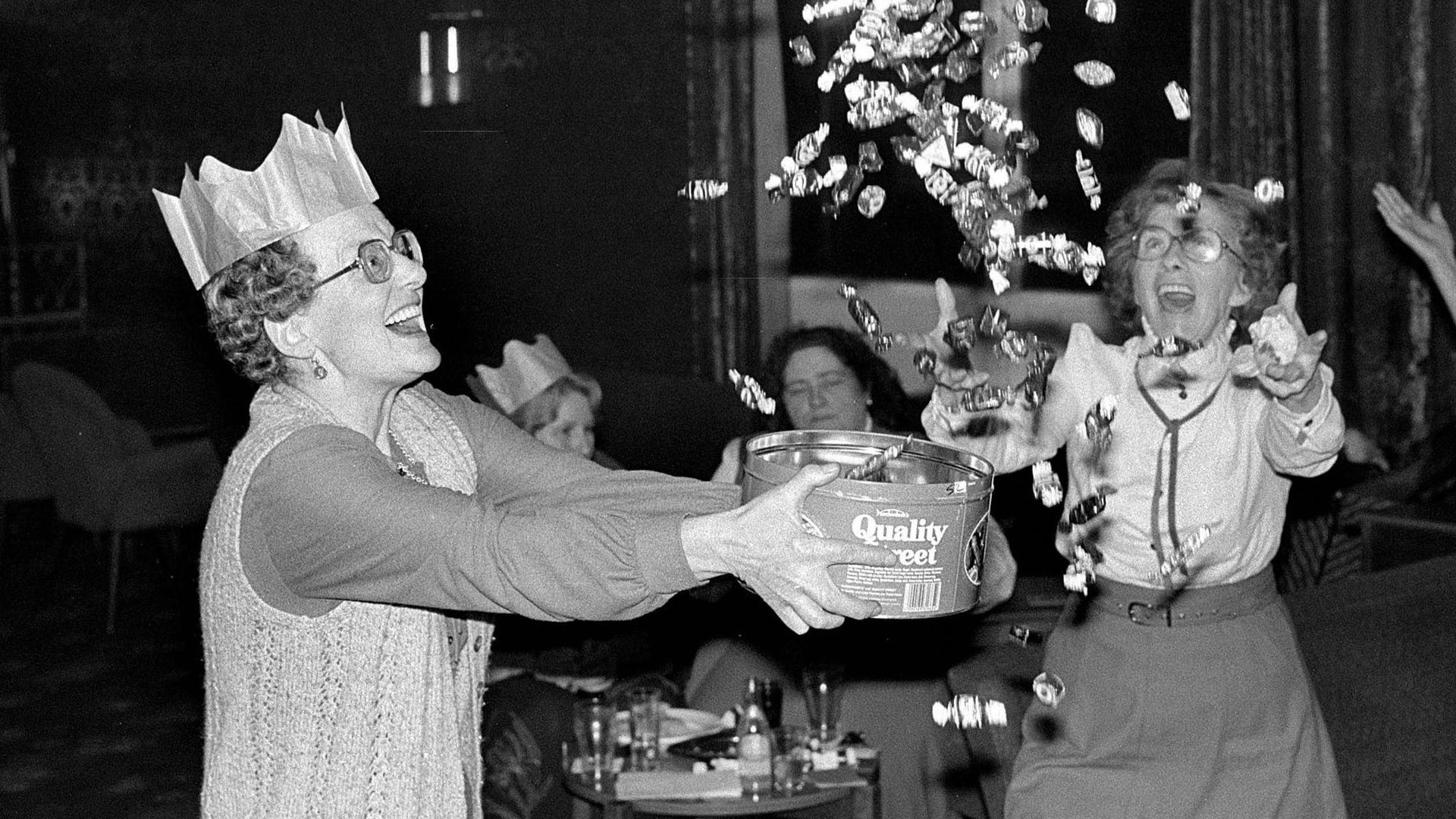 Two women, one of whom is wearing a party hat, look excited as the contents of a box of Quality Street are launched into the air.