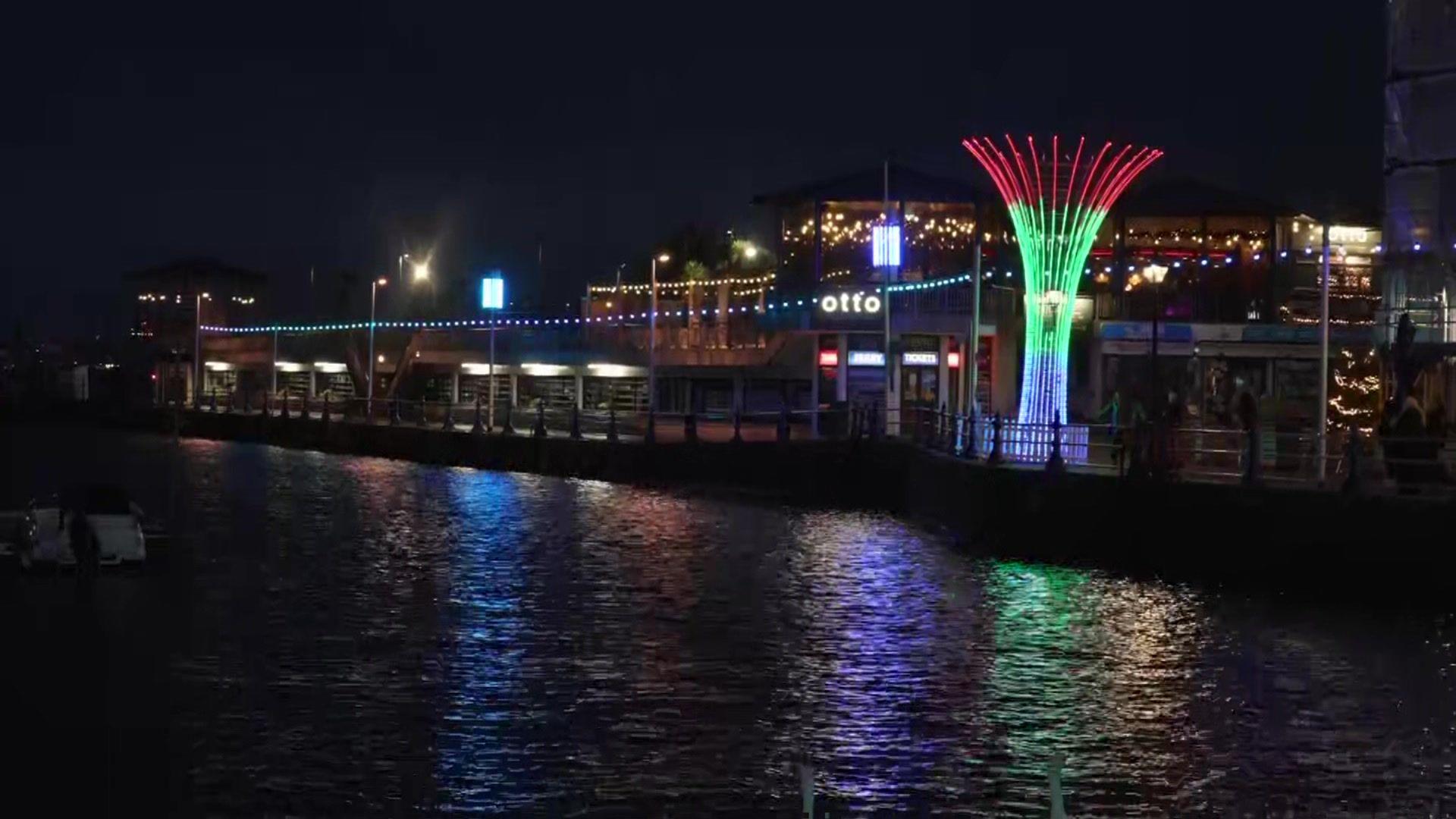 A body of water is seen in the foreground, with a string of lights and fountain of lights in blue, green, purple and red behind, against a backdrop of shops and eateries 