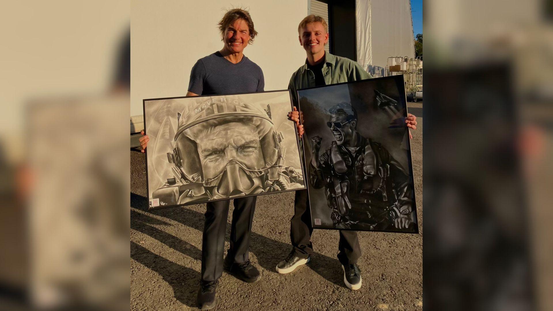 Actor Tom Cruise is standing with tattoo artist Wilf Elliott. They are each holding a large, framed black and white drawing of Tom Cruise. The actor is wearing a grey T-shirt and grey trousers, and the artist is wearing a green shirt and grey trousers. They are outside, standing on gravel with a large cream wall behind them.