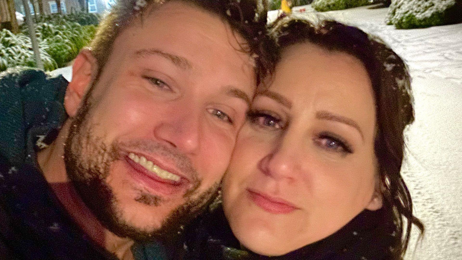 Chris and Carrie smile at the camera which their faces pressed together. It is snowing and they are clearly standing outside in the snow.