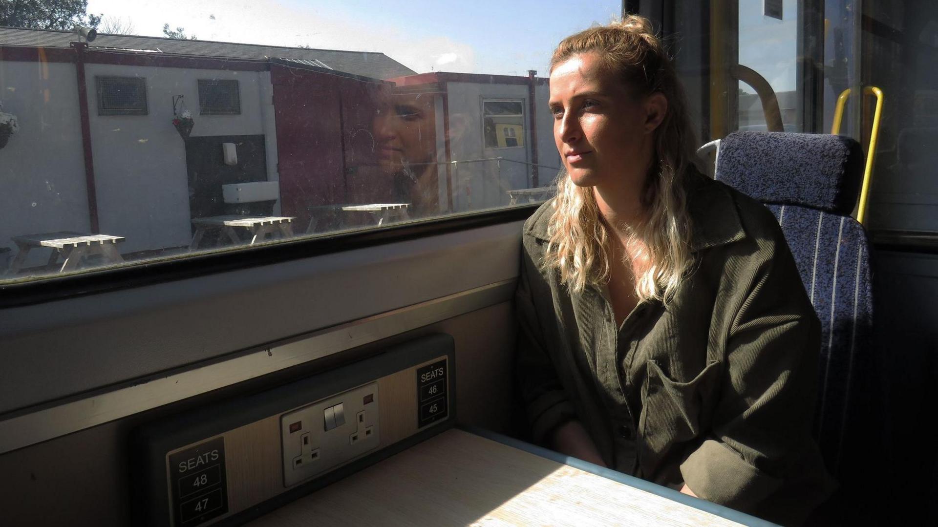 A woman sitting inside a train, looking out of the window as it goes past some industrial units, with her reflection in the glass.