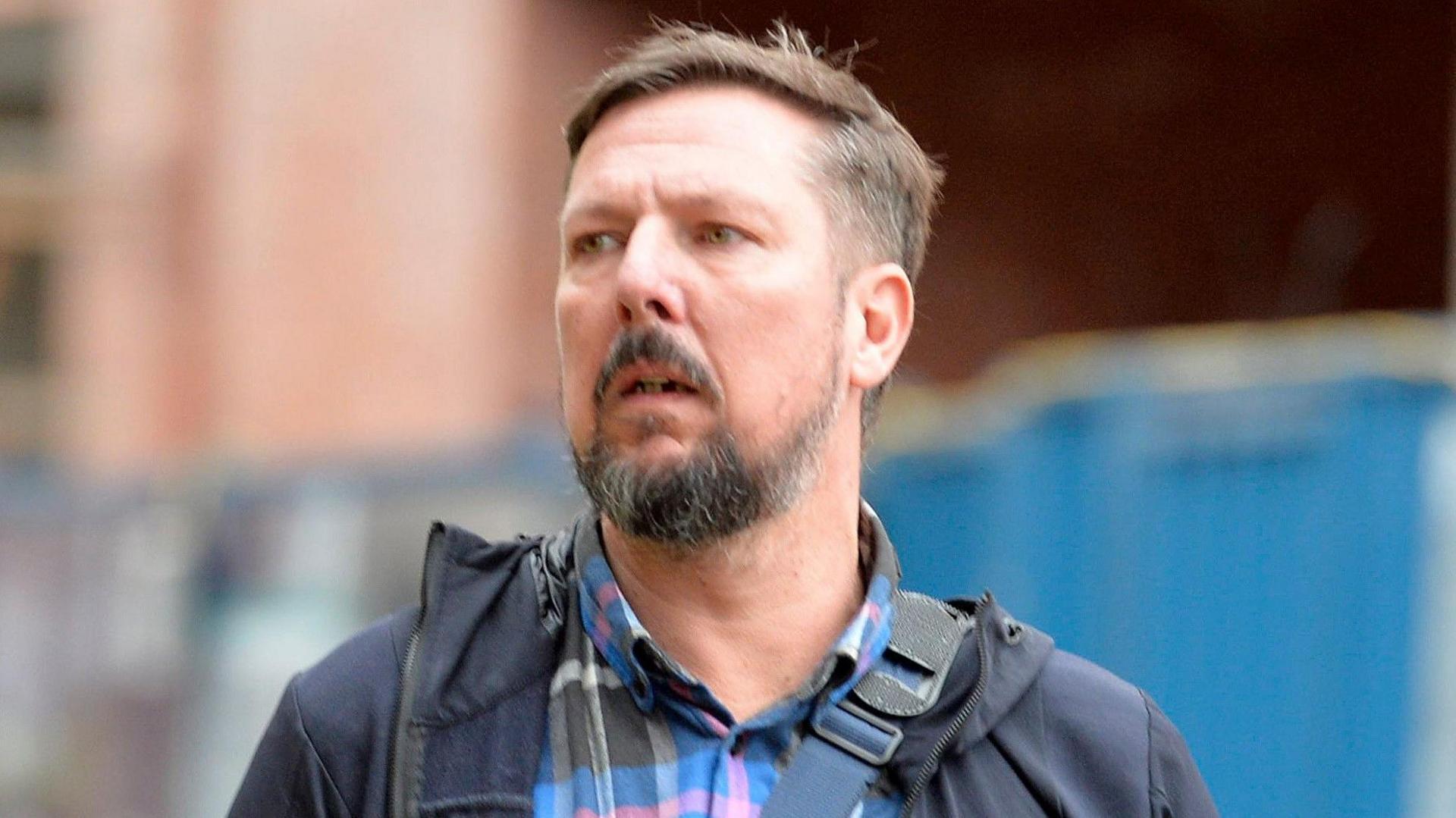 A man with short dark hear, beard and moustache, wearing a navy blue jacket, looking into the distance. 