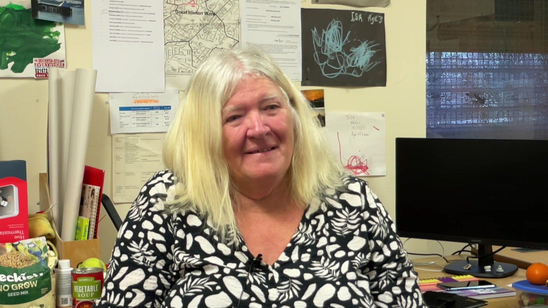 A woman in a black and white flowery top, sitting in an office, smiles at the camera.