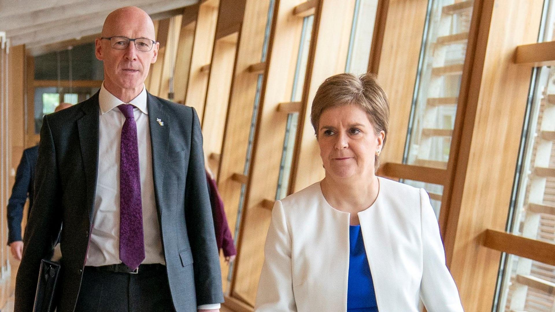 John Swinney and Nicola Sturgeon waslking down a long corridor at Holyrood. Swinney is on the left of frame in a dark suit and purple tie, carrying a folder. Sturgeon is on the right weating a white collarless jacket and electric blue blouse. 