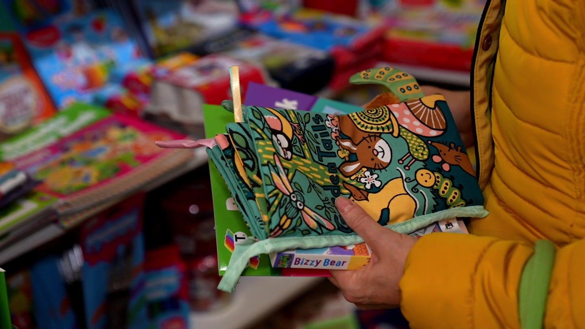 A woman in yellow coat hold three children's books in a shop. More books are in the background.