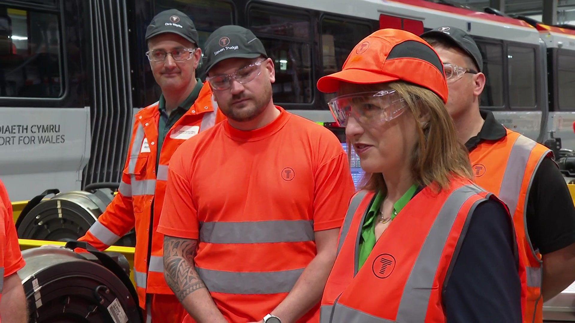 Rachel Reeves wearing orange hi-viz to the right of the photograph, surrounded by workers a train depot to her left. All are wearing protective eye wear.