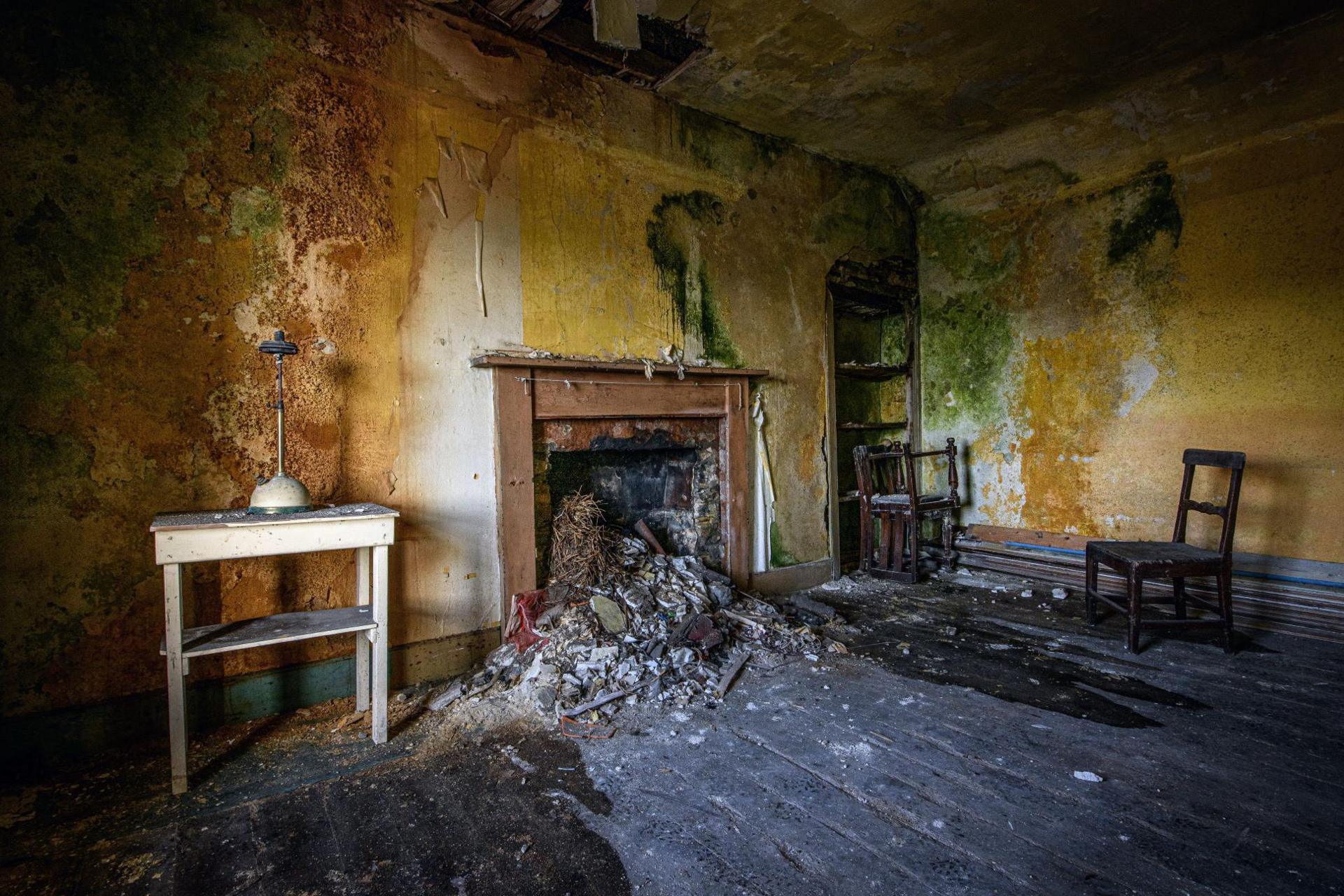 The room has bare floorboards and the walls are stripped of wallpaper and covered in mould. There are two wooden chairs along one wall and opposite them a small white table. Debris spills out from a fireplace. 