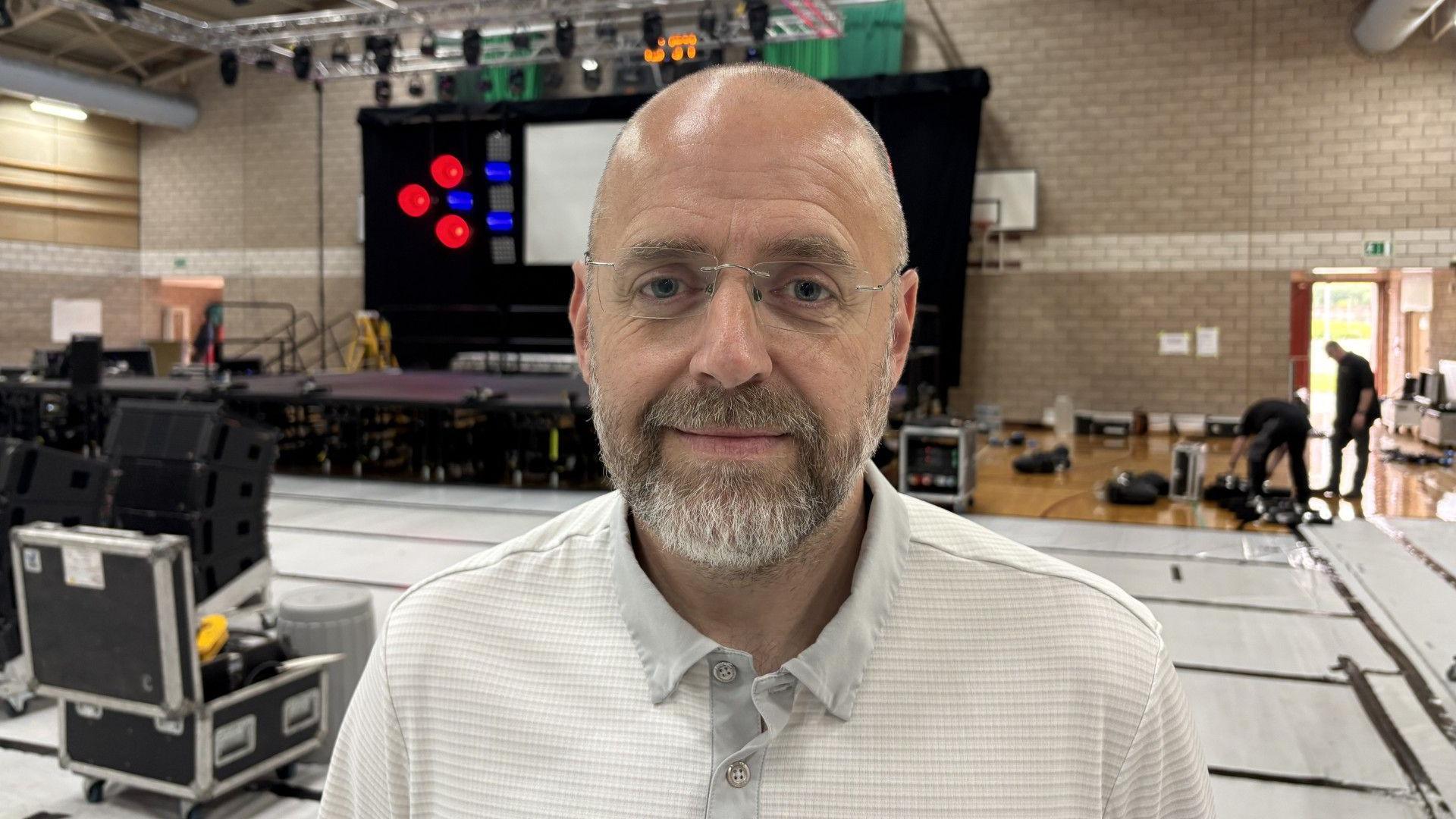 A bald man with facial hair and glasses. He is looking at the camera and standing in a sports hall covered in musical equipment.