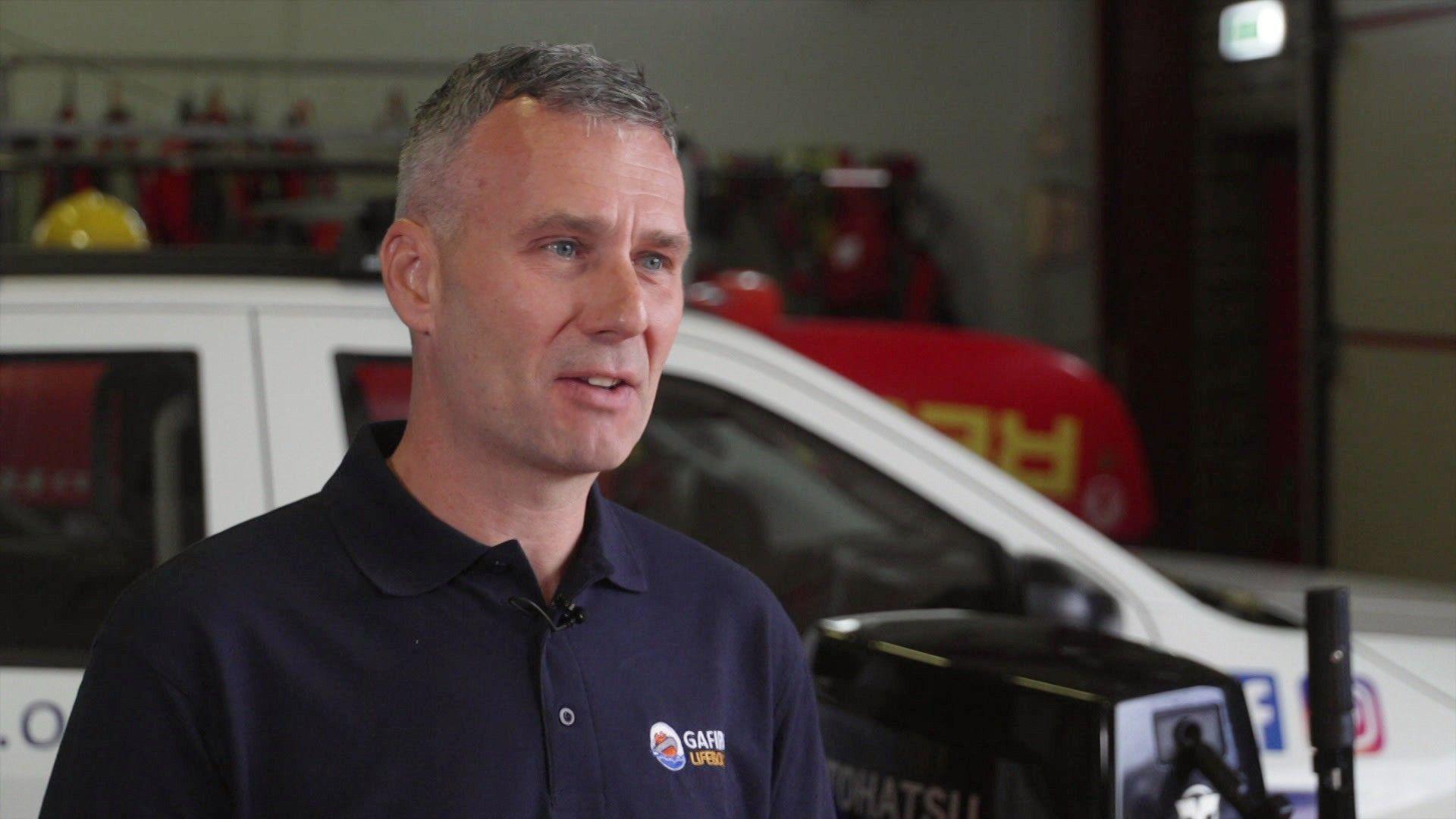 James Baggott gives a TV interview inside a lifeboat station. He has short, grey hair and wears a dark polo shift with the lifeboat station badge on the front.