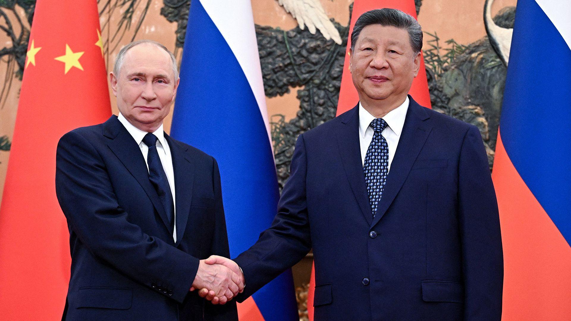 Vladimir Putin stands next to Xi Jinping while shaking his hand and both look solemnly towards the camera wearing suit and ties in front of Russian and Chinese flags, while the huge artwork adorning the Great Hall of the People can just be seen behind the flags.