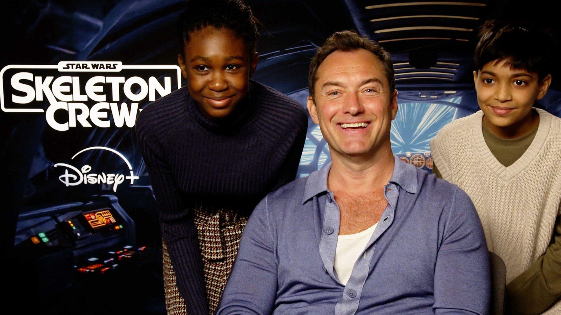 A girl and boy smile with actor Jude Law, in front of a background that says Star Wars Skeleton Crew