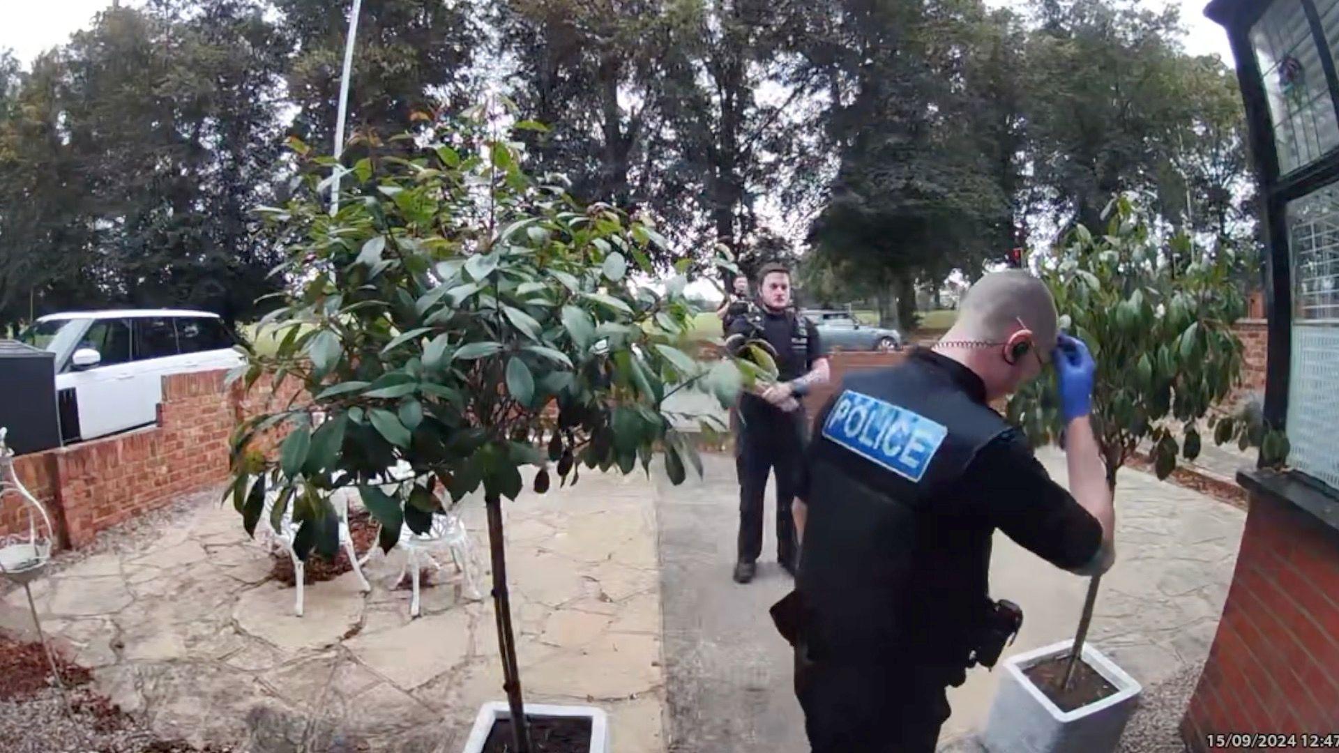 Police officers appear at the door of Maria Botterill's house in Northampton. At least three male officers are visible. They are standing in the driveway, which is enclosed by a wall. There are large trees in the background