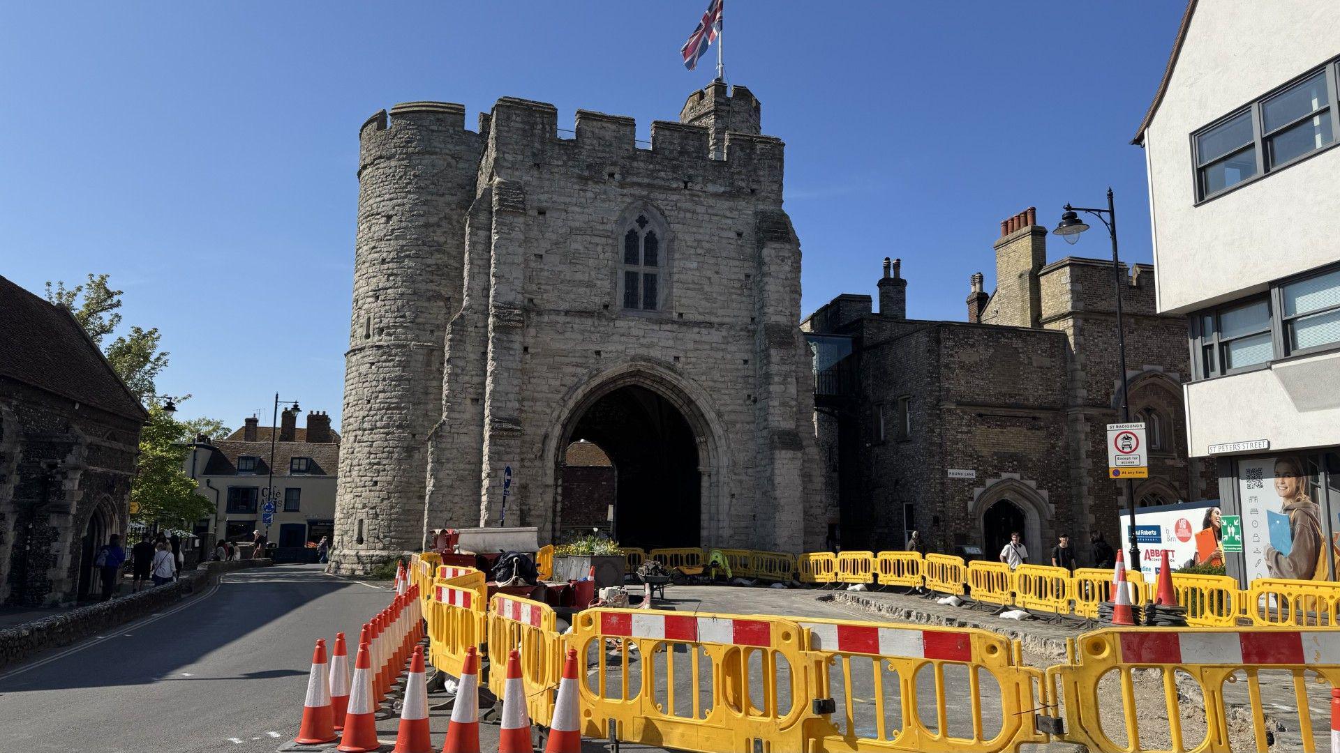 A stone tower with a large archway underneath it, flying a union jack flag from its turrets. Road access to the archway is blocked by orange traffic cones and yellow plastic barriers.