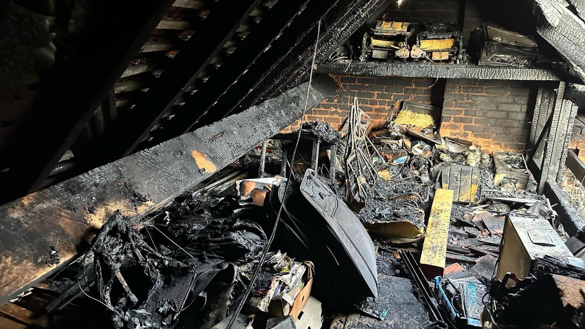 A charred loft space, with tangles of wire, objects and planks all blackened and melted together by fire.