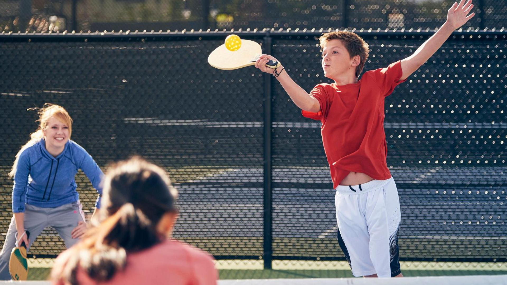 Young people playing pickleball. A boy hits a ball with a wooden bat. He wears a red t-shirt and white shorts.