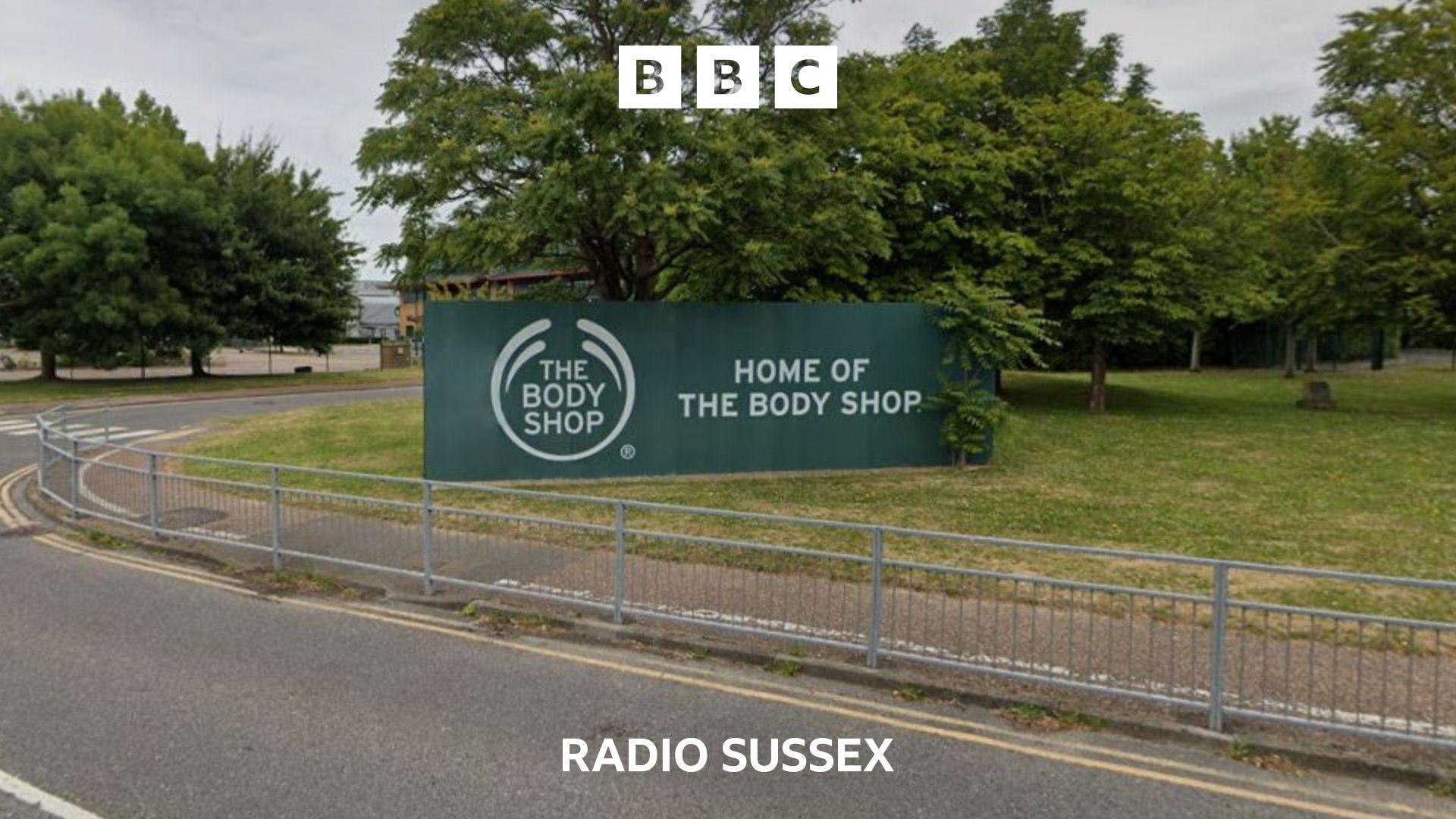 A large green and white The Body Shop sign sits on grass. There is a road and pavement in front, separated by a metal fence.