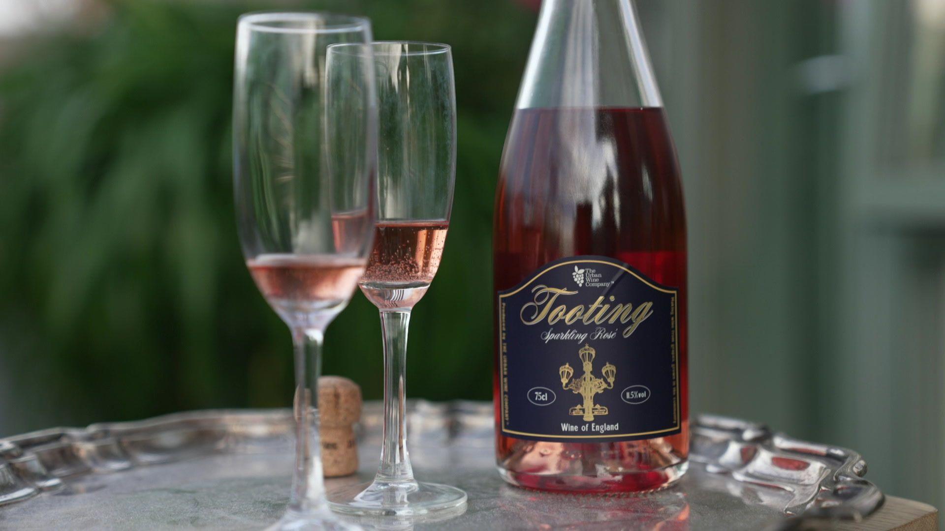 An open bottle of Tooting Sparkling Rosé on a silver tray. Next to it are two glasses with varying levels of wine in each glass, and a cork from the bottle.