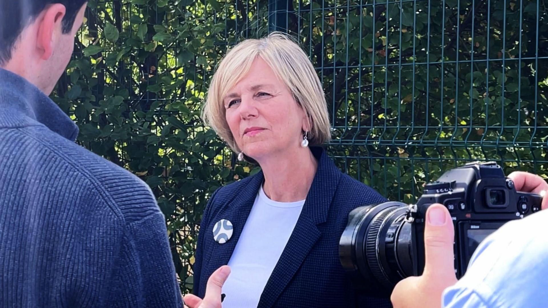 A woman with cropped blonde hair, wearing a blue blazer and white top, looking at a man wearing a blue jumper. An SLR camera is also pointing at her. Behind her is a metal fence with bushes growing behind it.