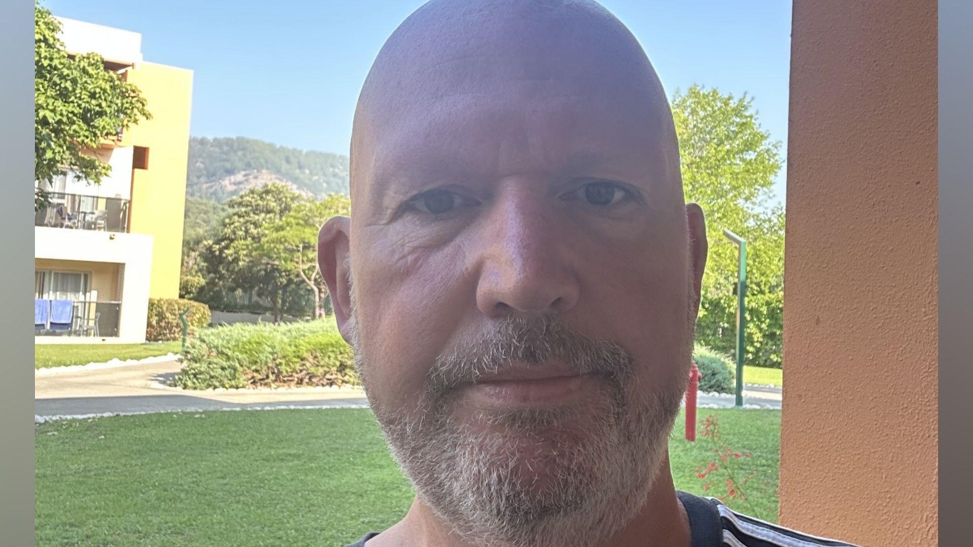 Gavin Sparks takes a selfie at his hotel. He has a bald head and a short grey beard. He is wearing a black Adidas t-shirt with white stripes on his arm.