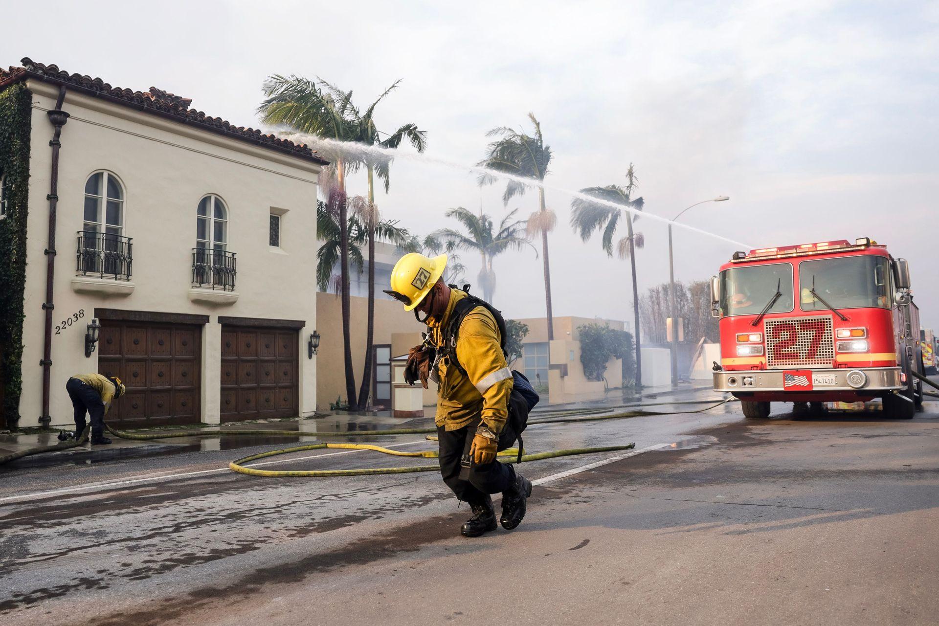 A Los Angeles County firefighter drags a water hose in front of a fire engine that's shooting water at a building, as they take up positions to battle the Palisades wildfire in Malibu on Wednesday.