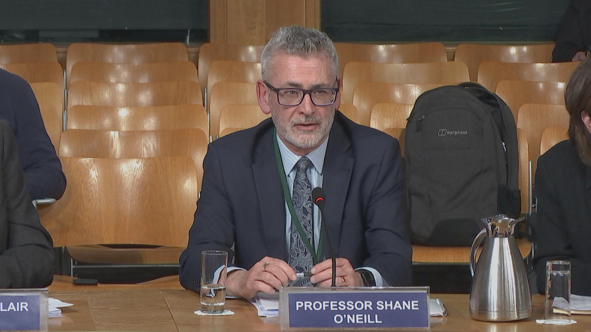 Professor Shane O'Neill, a man with grey hair and glasses, sits at a committee table