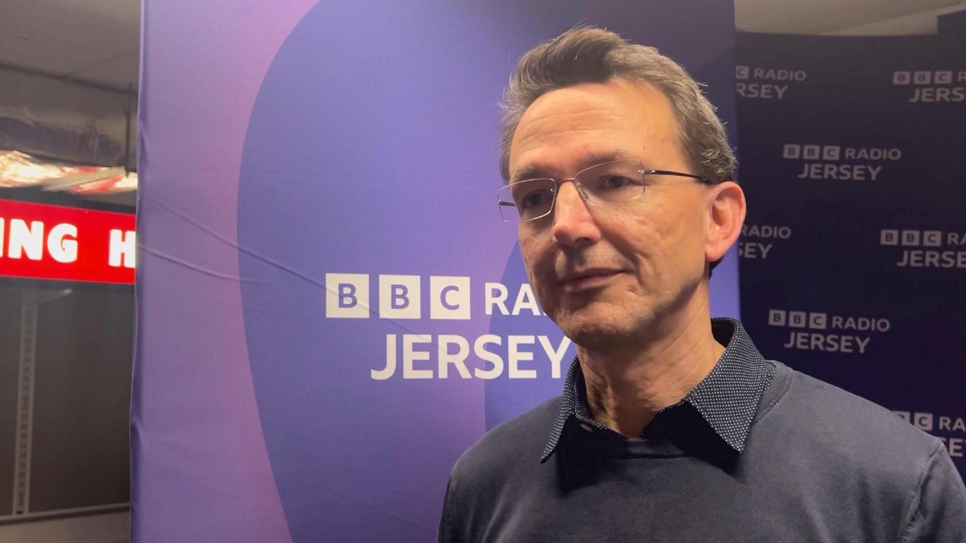 Kirsten Morel - a man wearing rimless rectangle glasses, a navy collared shirt and a navy jumper on top. Behind is the purple BBC Radio Jersey banner.