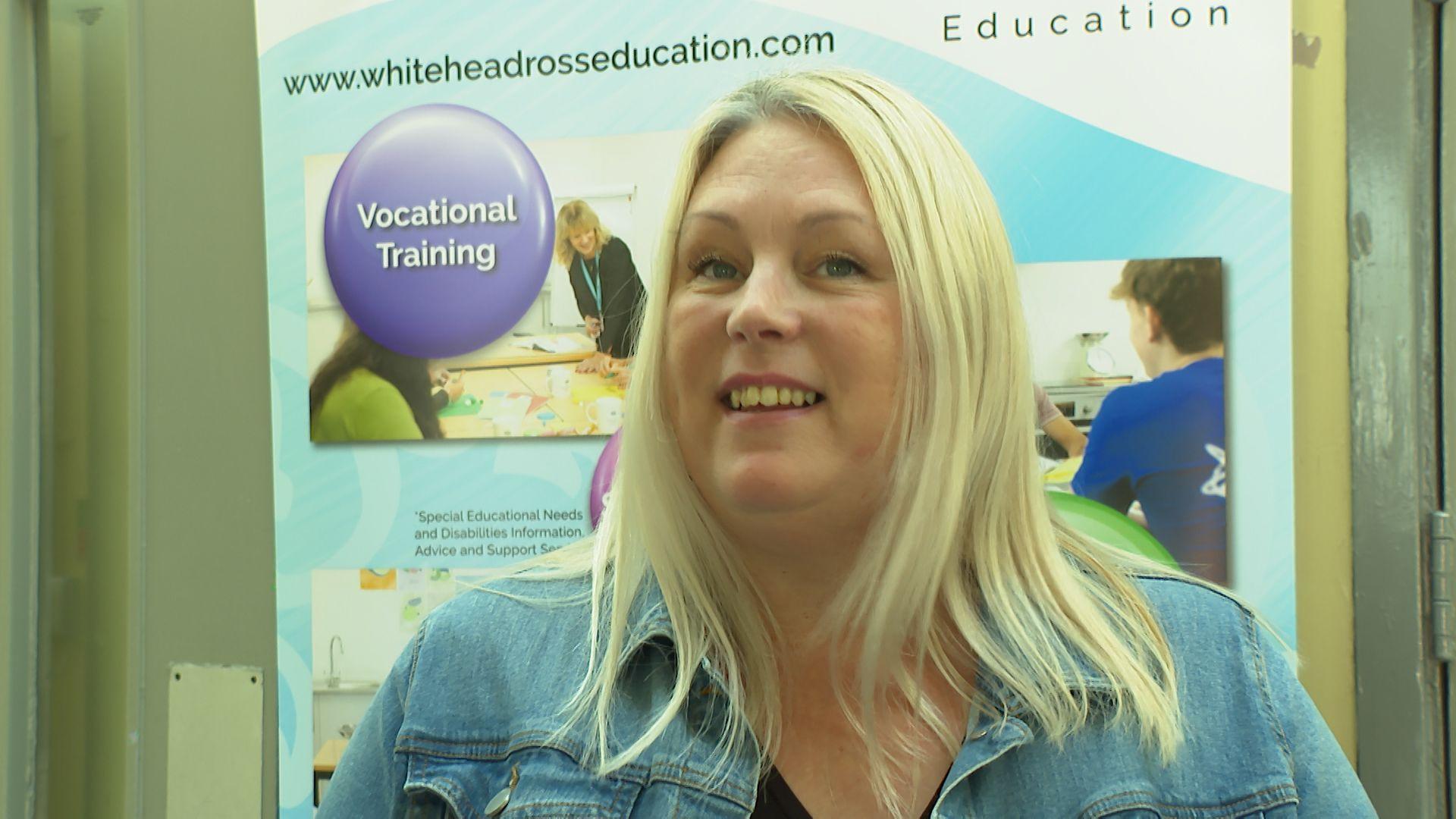 Menyw  yn sefyll o flaen baner Whitehead-Ross Education. Mae hi'n gwisgo siaced ddenim ac mae ganddo wallt golau.