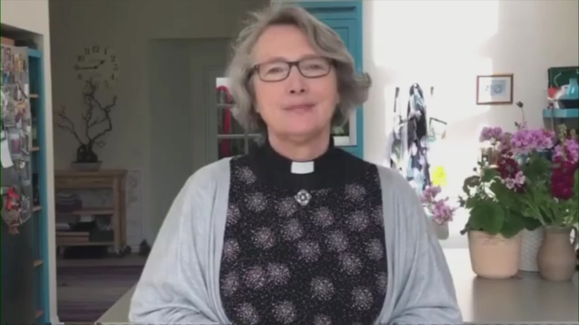 Sue Colman is wearing a dog collar and a grey jumper and grey and black top. She has grey, shoulder length hair and dark rimmed glasses. She is apparently standing in a kitchen, with pot plants to her right