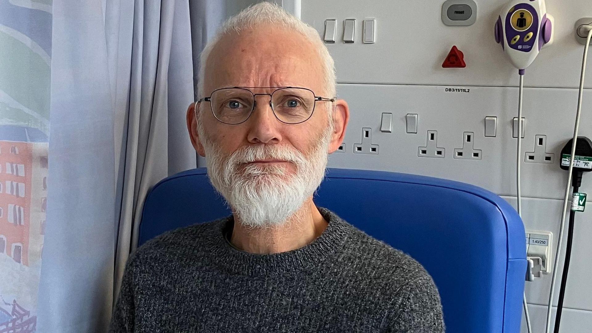 A man with grey hair and beard. He is wearing a grey jumper and sitting in a blue hospital chair