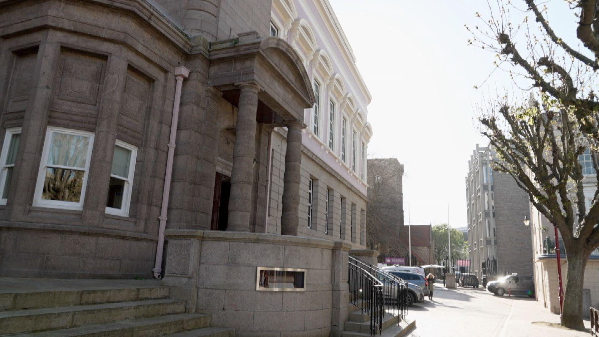 A stone building with a number of windows. A path runs in front of the building. Cars are parked at the entrance. Small steps lead up to the entrance of the building.