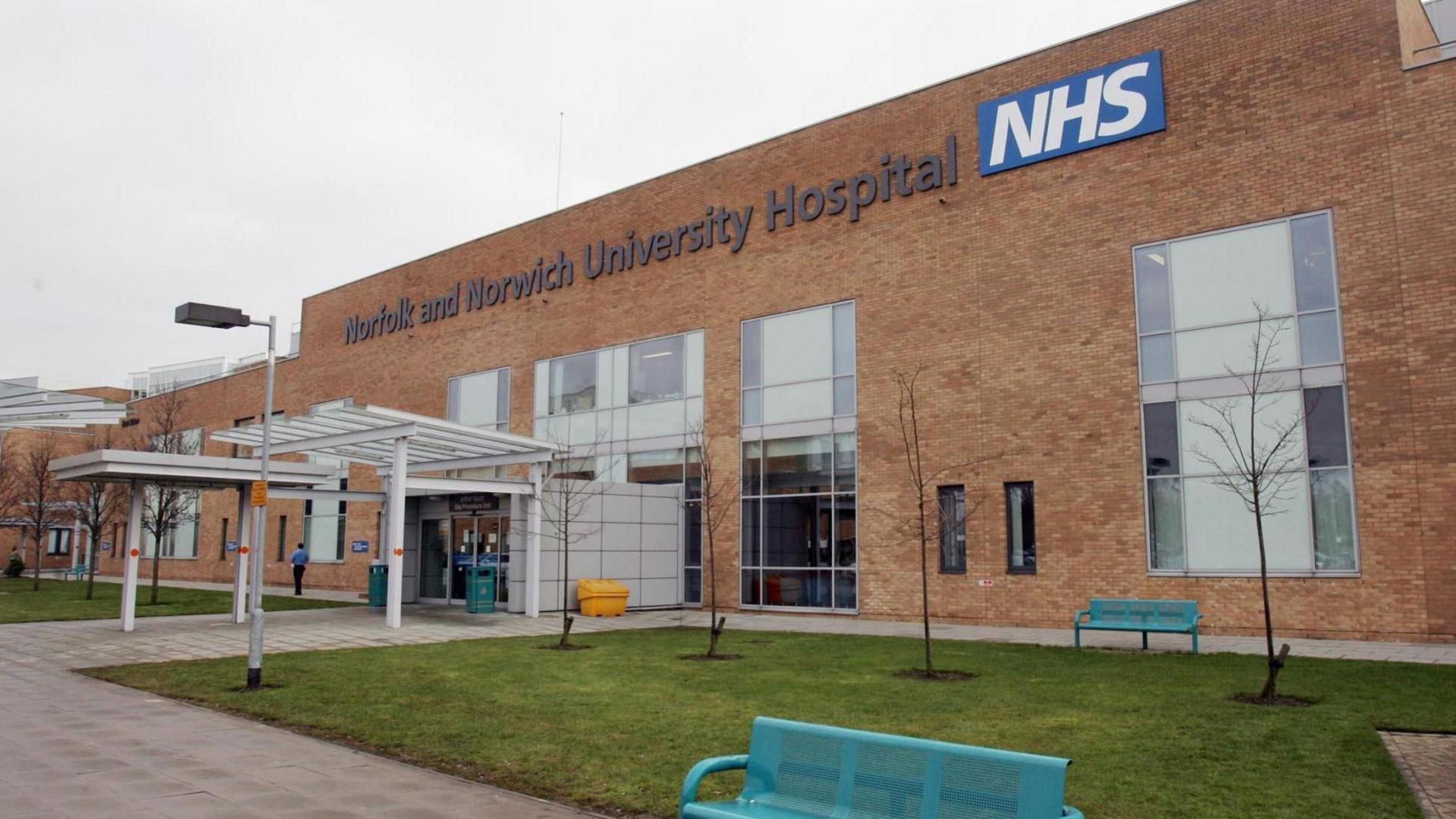 The entrance to the Norfolk and Norwich University Hospital. It is a light brown brick building with a white entrance canopy. Two turquoise benches are located by a patch of grass.