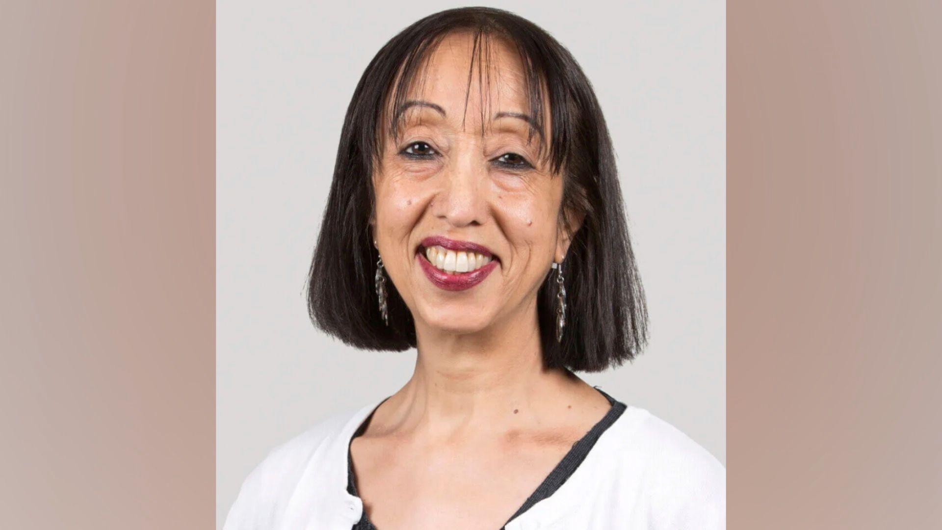 Nina Ali has straight dark hair down to her neckline. She is smiling at the camera and has dark red lipstick on. She is wearing a white cardigan with black top underneath, and has long earrings on as well.