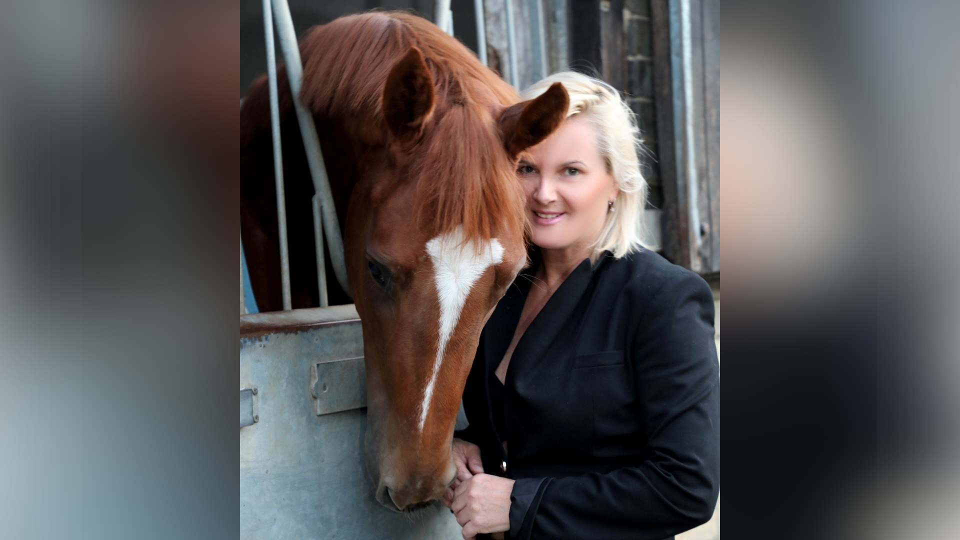 A picture of Gay Kelleway with a chestnut coloured horses. The horse has its head over the stable door and is looking down. Gay is smiling at the camera. She has short blonde hair and it wearing a black suit.