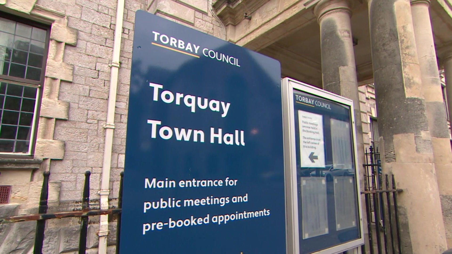 A blue sign with has the words 'Torbay Council' at the top. Below it there is white writing which says 'Torquay Town Hall'  and 'Main entrance for public meetings and pre-booked appointments'. Behind the sign is a building with a window on the left and bollards on the right. 