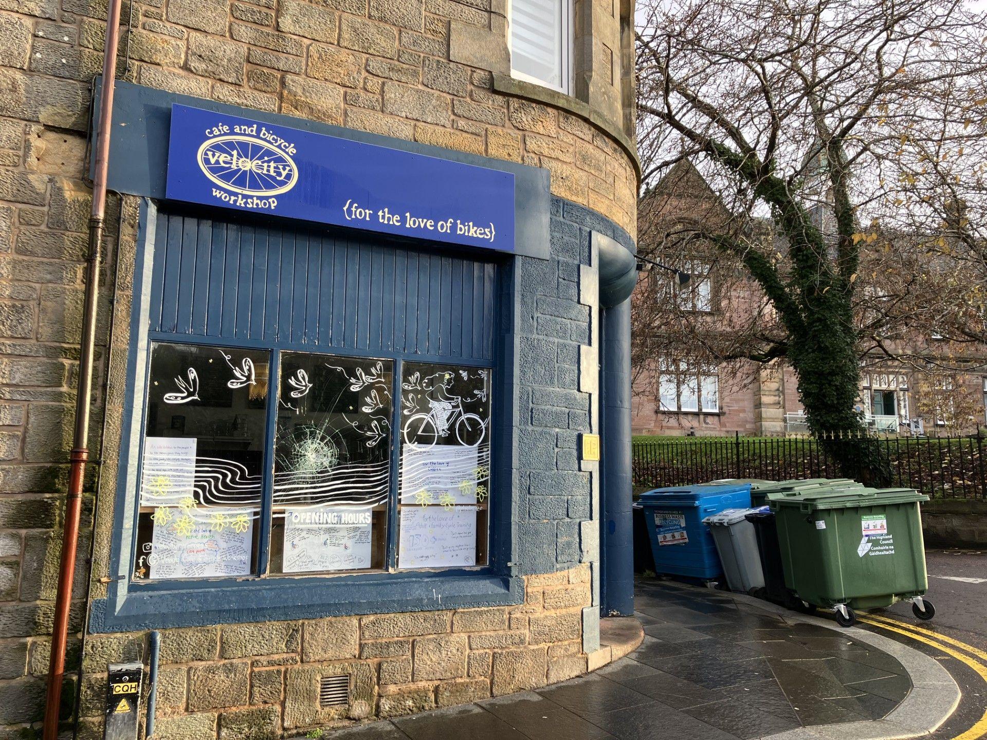 The exterior of the business has a blue frontage. A sign above a window includes the words: "For the love of bikes". There are industrial wheelie bins on a corner of the street next to the building.