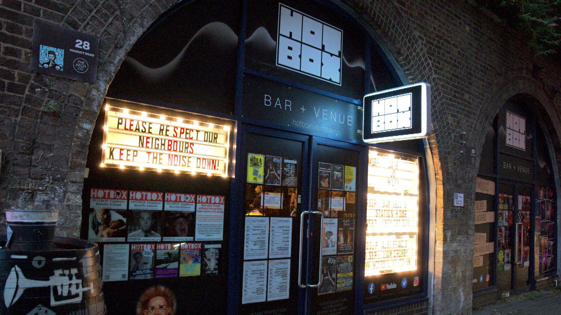 The entrance of a small venue. There are signs on the door that say "Hot Box", and lots of posters.