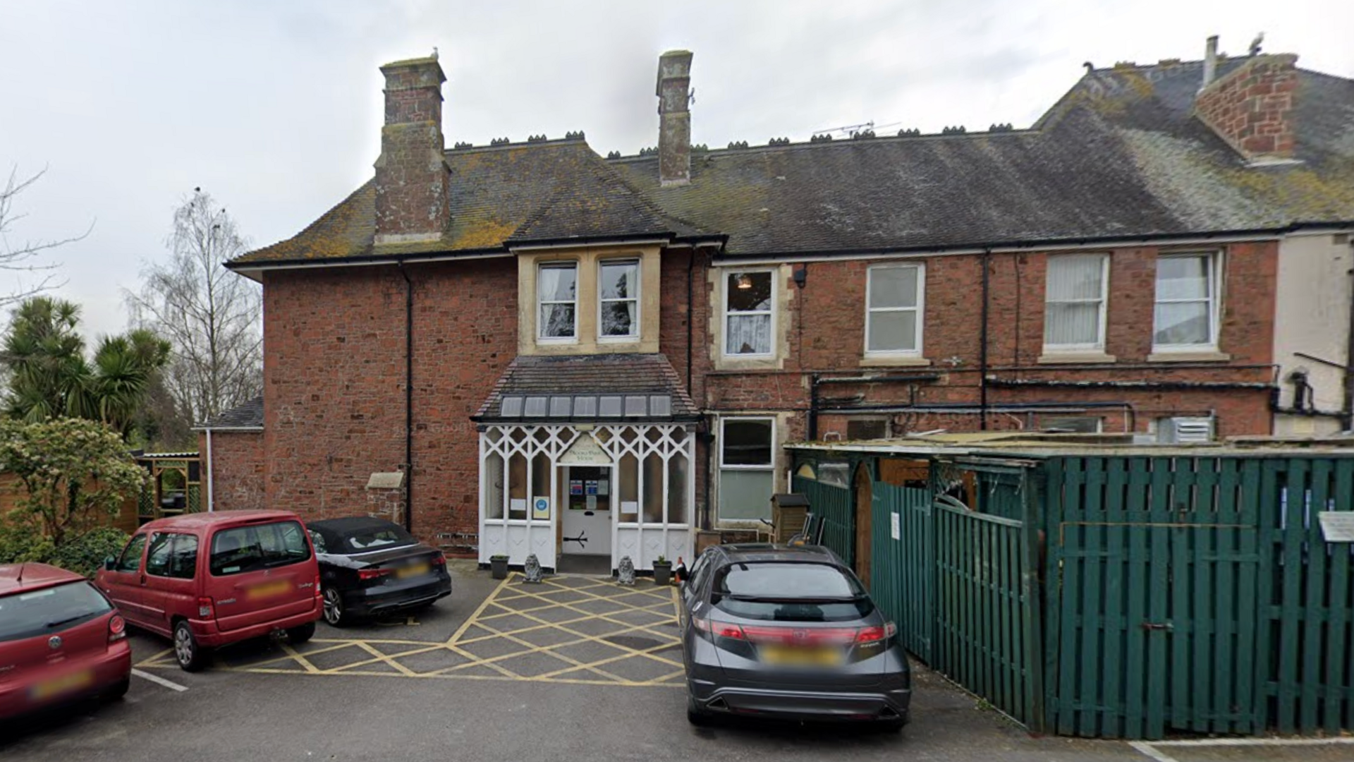 A brick two storey building with a grey slated roof with three chimneys and a white porch, two red cars and two black cars parked outside on a grey sky day.