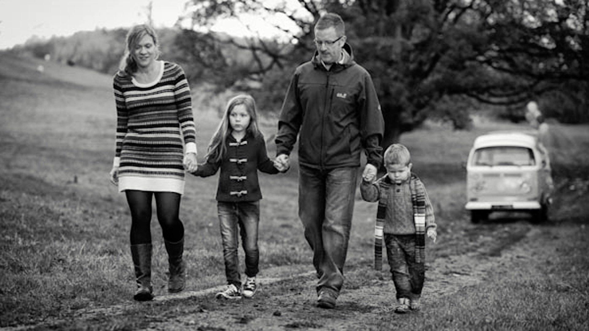 A black and white photograph of the Sherriff family, in coats and scarves, holding hands as they walk along a path cut through a muddy field, with a VW camper van in the background