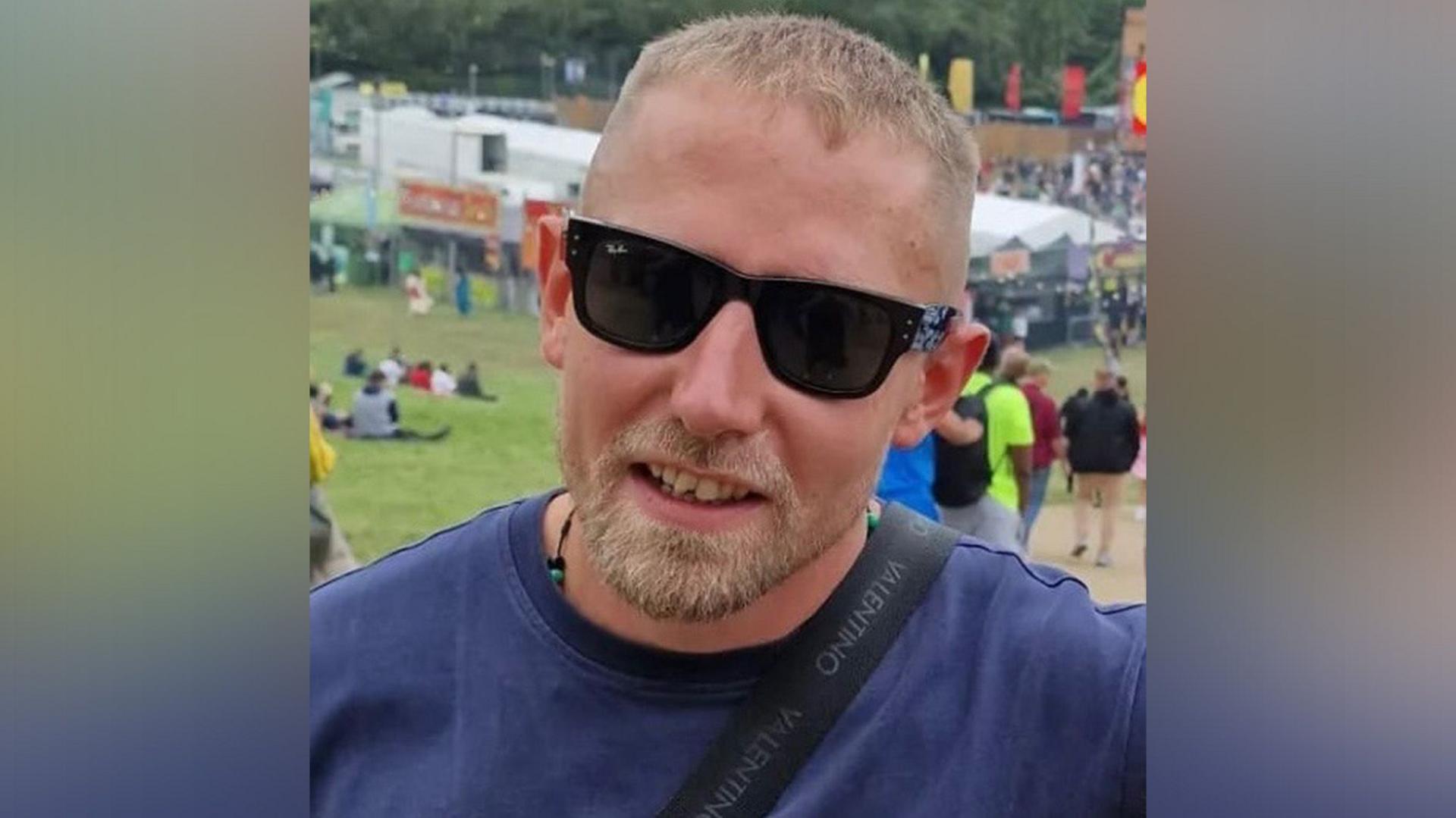 Ben Hardy on a summer's day outside, wearing a blue T-shirt and sunglasses, with a strap over his shoulder. He has short hair and a beard.