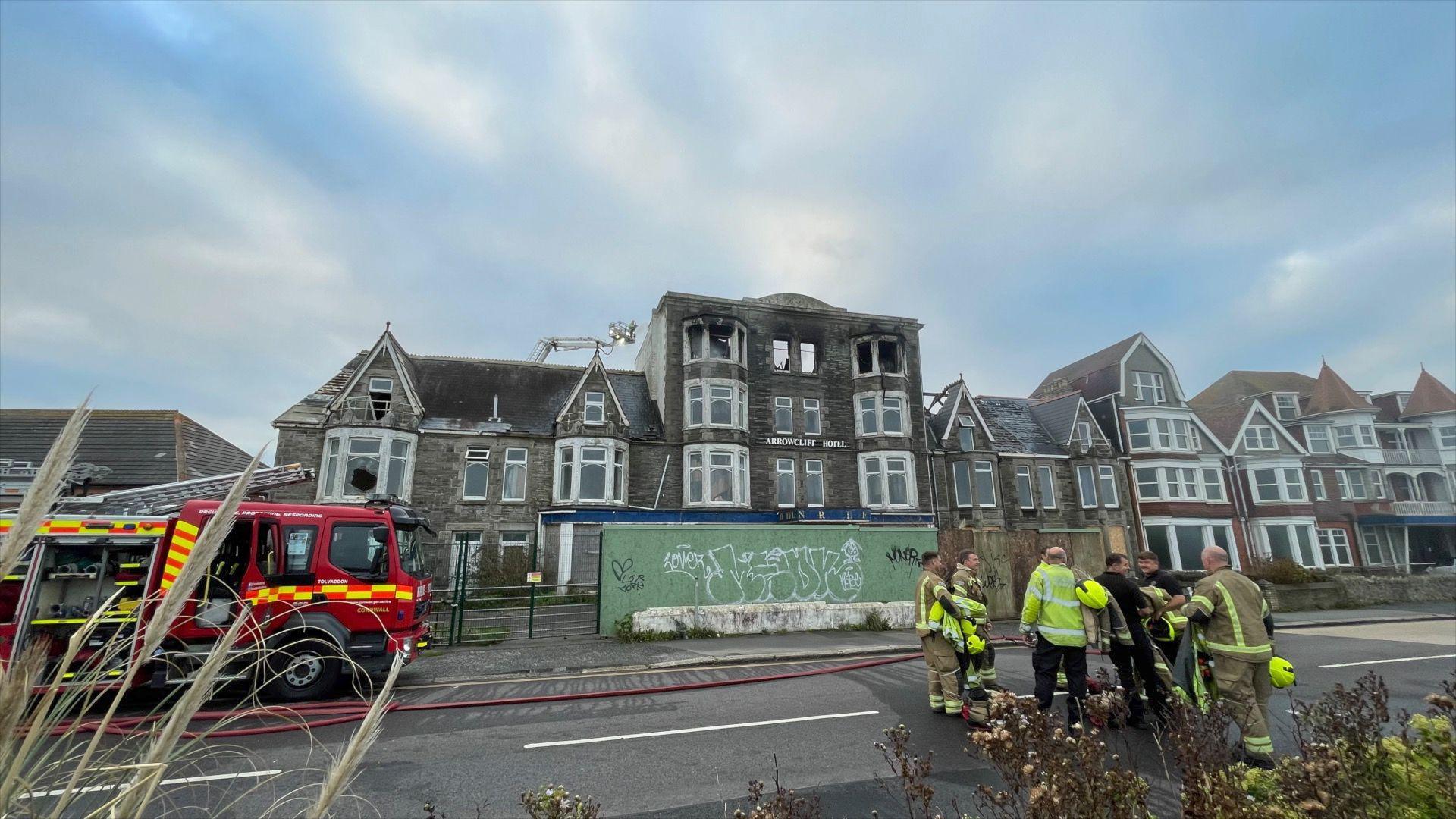 The Narrowcliff Hotel seen with scorch marks at the windows of its upper floor. A fire engine is parked outside and there are about half a dozen firefighters stood outside in a group wearing their uniform.