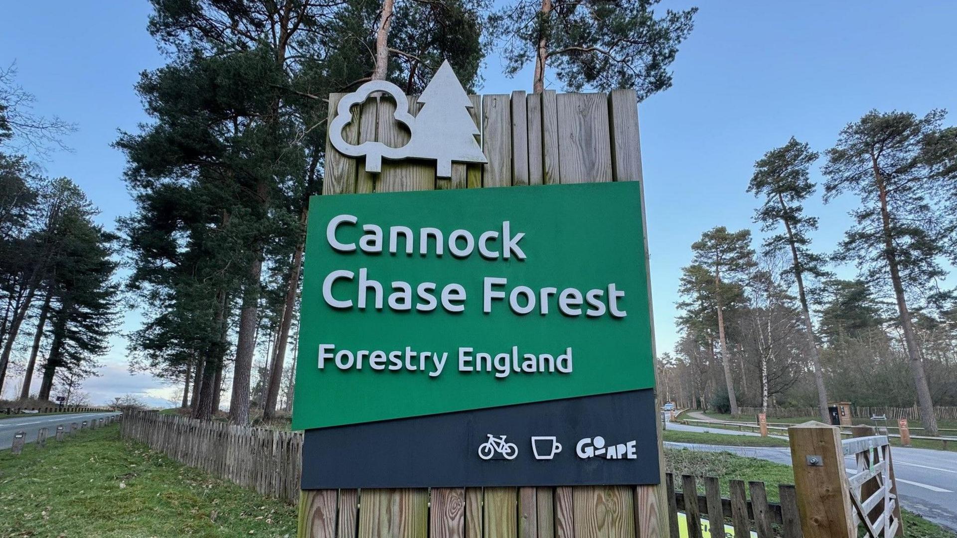 A sign at the entrance to Cannock Chase Forest, with tall trees visible behind. The sign has a green background with white lettering.