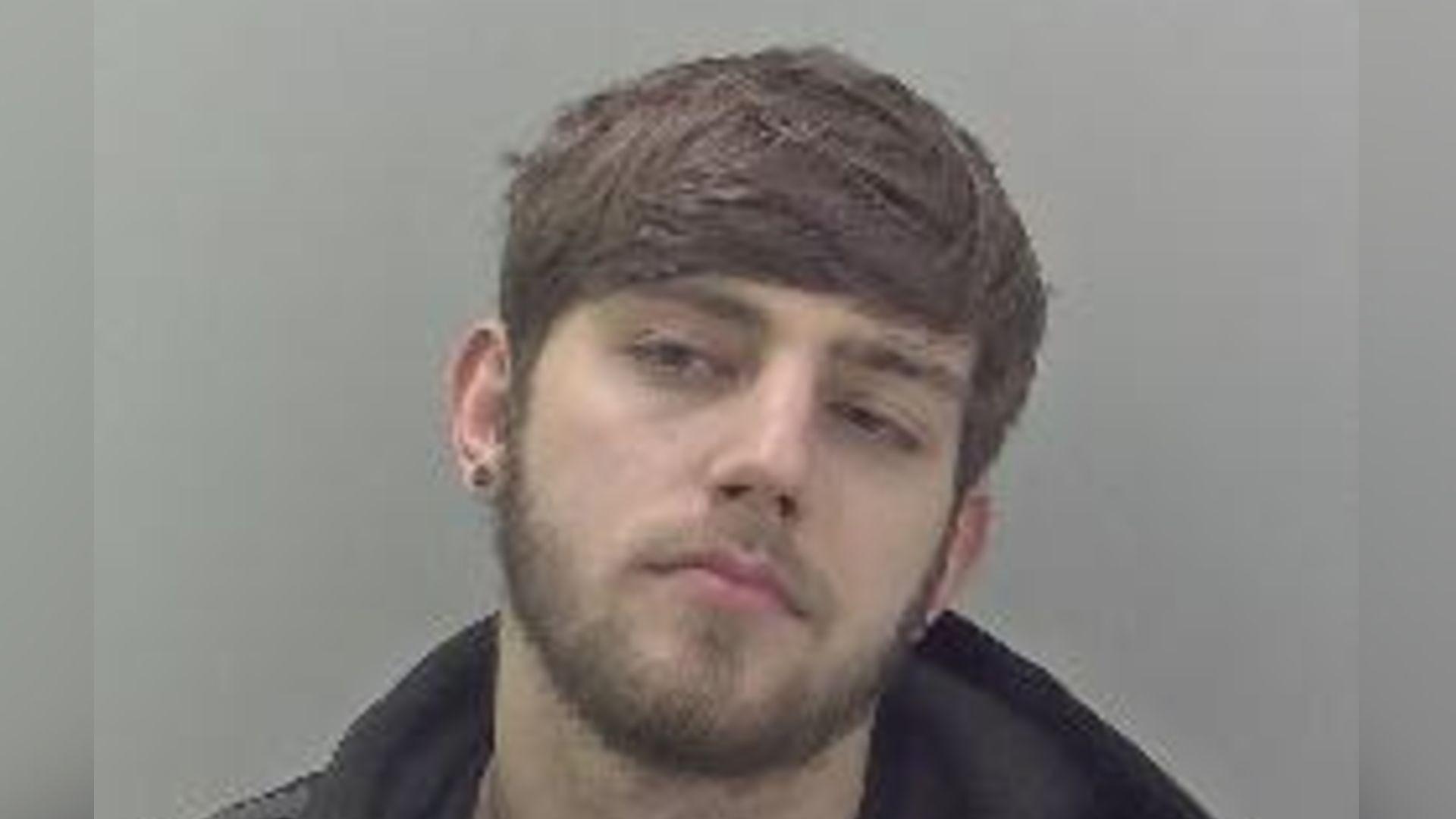 A police photograph of paedophile Scott Chapman. He is waring an earring, has brown hair and a beard. He is refusing to look straight into the camera, tilting his head. He is also wearing a dark jumper and hoodie. 