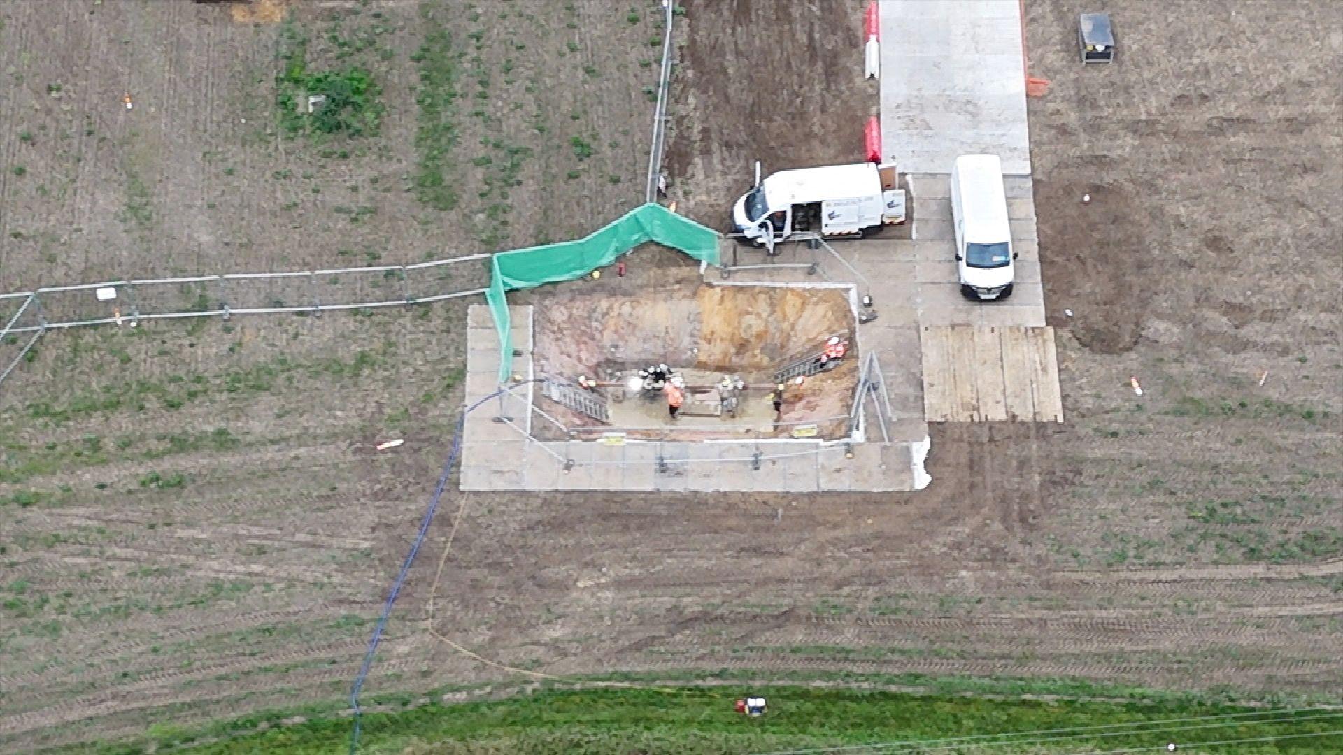 A drone image of a field, showing two parked white vans and a fenced off rectangular area in which a smaller rectangular pit has been dug, approximately 20 to 30 ft deep. Several workman in hard hats and hi-vis jackets are in the pit.