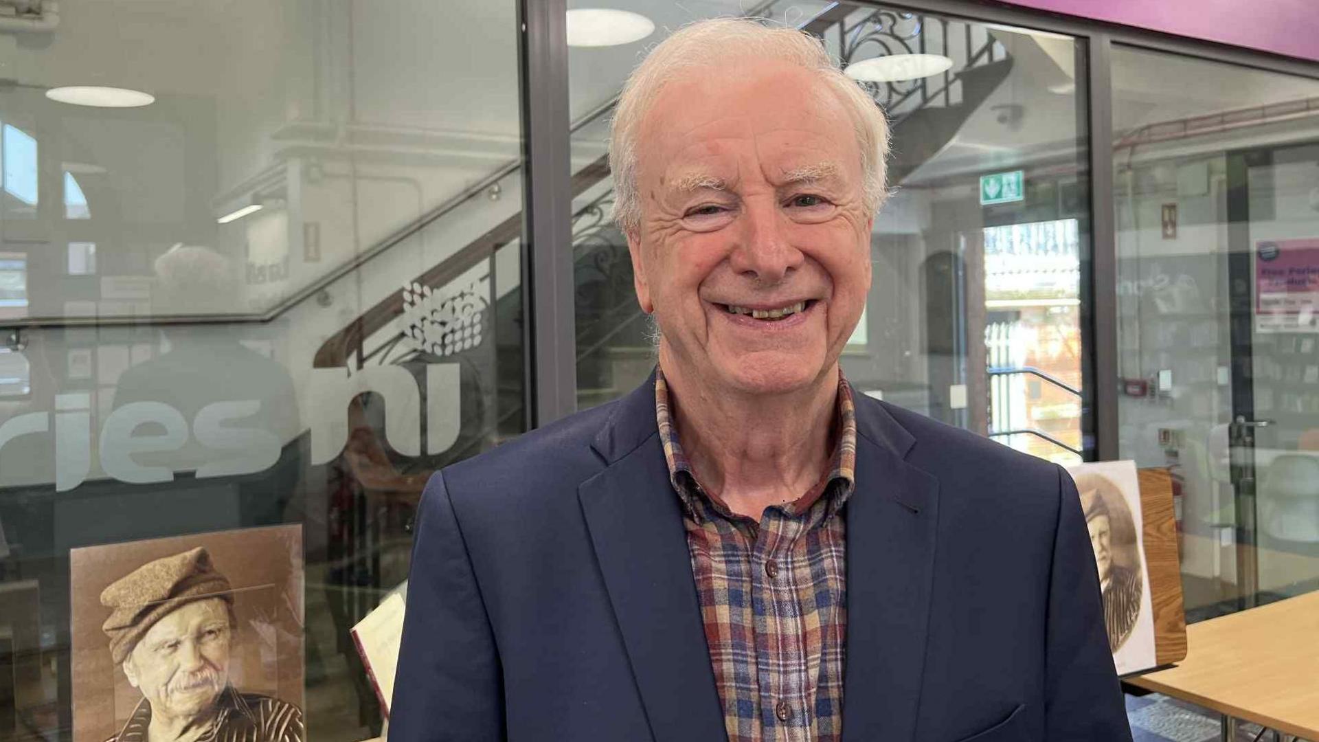 Seamus McKee smiles at the camera, he has short white hair and is wearing a dark blue suit jacket over a checked shirt. He is standing in front of a glass partition in Falls Road Library.